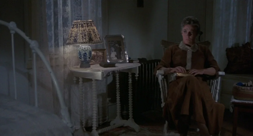 The Other (1972) movie still: medium — A dimly lit room contains a white metal bed frame and a side table. An elderly woman sits in a rocking chair…