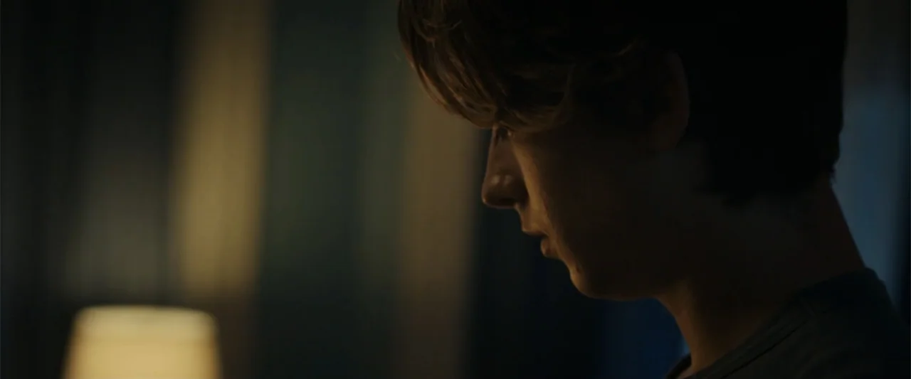 Chemical Hearts (2020) movie still: closeup — A dark, dimly lit bedroom at night with a blurred lamp in the background. A young man sits with his…