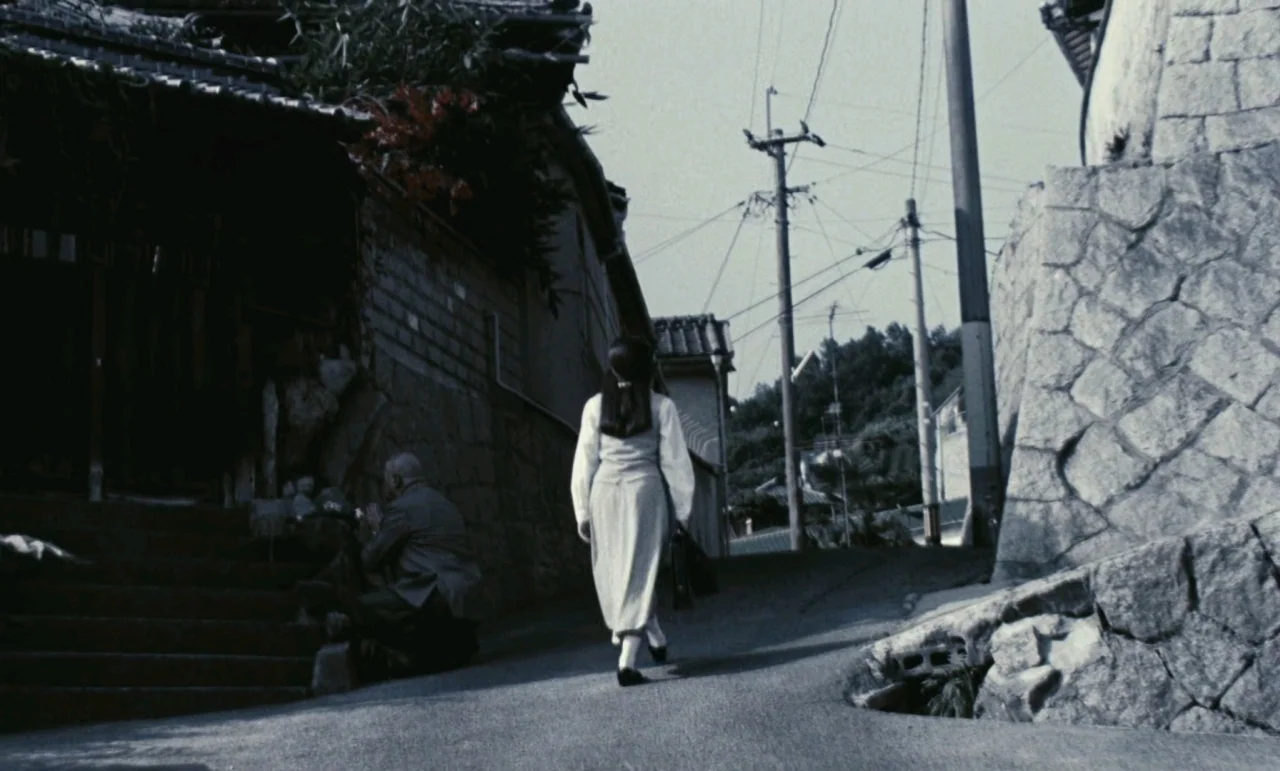 Chizuko’s Younger Sister (1991) movie still: fullbody — A narrow uphill street is flanked by traditional stone walls and wooden buildings. A woman…