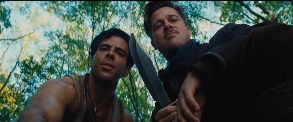 A dense green woodland setting with sunlight filtering through the trees. Two men in period military-style clothing look downward toward the ground.