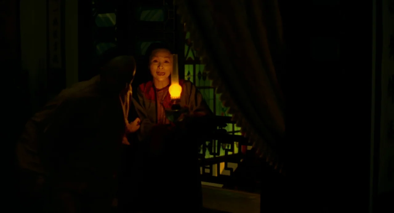 Flowers of Shanghai (1998) movie still: medium — A dimly lit room with ornate dark wood fretwork visible in the background. An elderly woman stands…
