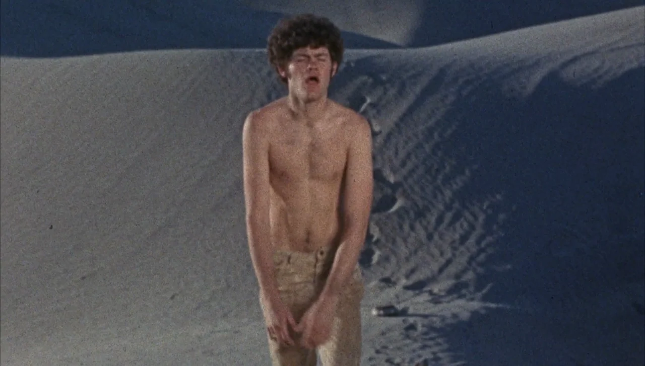 Head (1968) movie still: medium — A vast sandy desert at night with undulating dunes. A shirtless young man with curly hair stands alone in the sand.