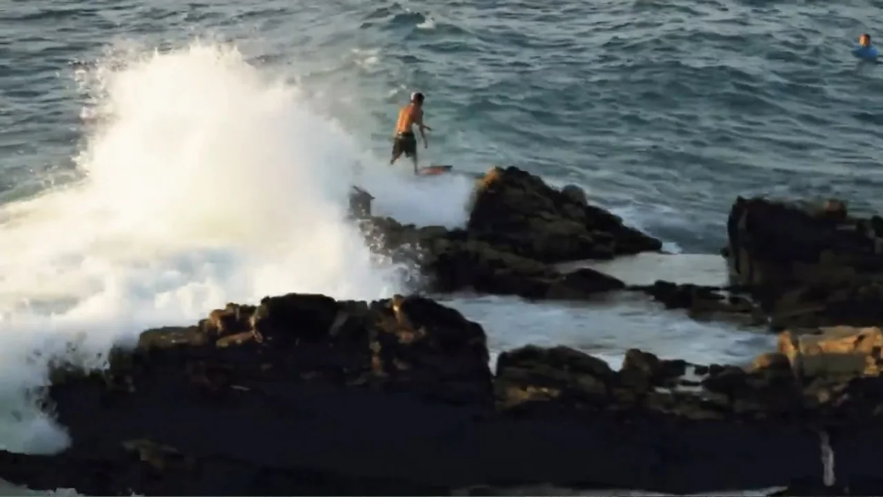 El sueño (2020) movie still: wide — A rocky shoreline is pounded by incoming waves during the late afternoon. A lone surfer stands on his board…