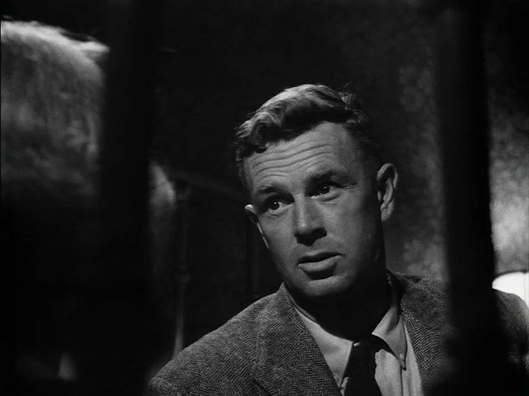 The Killing (1956) movie still: closeup — A dimly lit room with textured wallpaper and foreground vertical obstructions framing the shot. A man in a…
