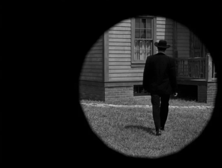 The Night Of The Hunter (1955) movie still: fullbody — A man walks across a lawn toward the side of a wooden house. The scene is framed through a…