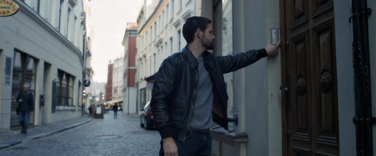 The Good Neighbor (2022) movie still: medium — A man in a leather jacket stands on a European-style cobblestone street. He reaches his hand out to…