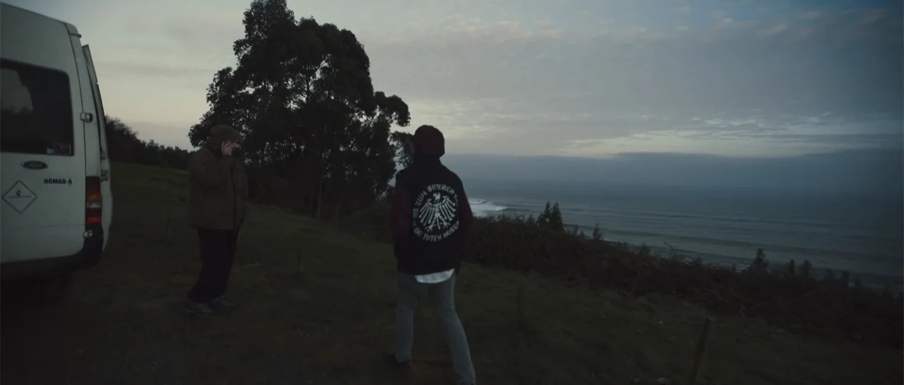 Taking Back the Waves (2017) movie still: wide — A coastal cliff overlooks a dark ocean under an overcast sky during dusk. One person in a dark vest…