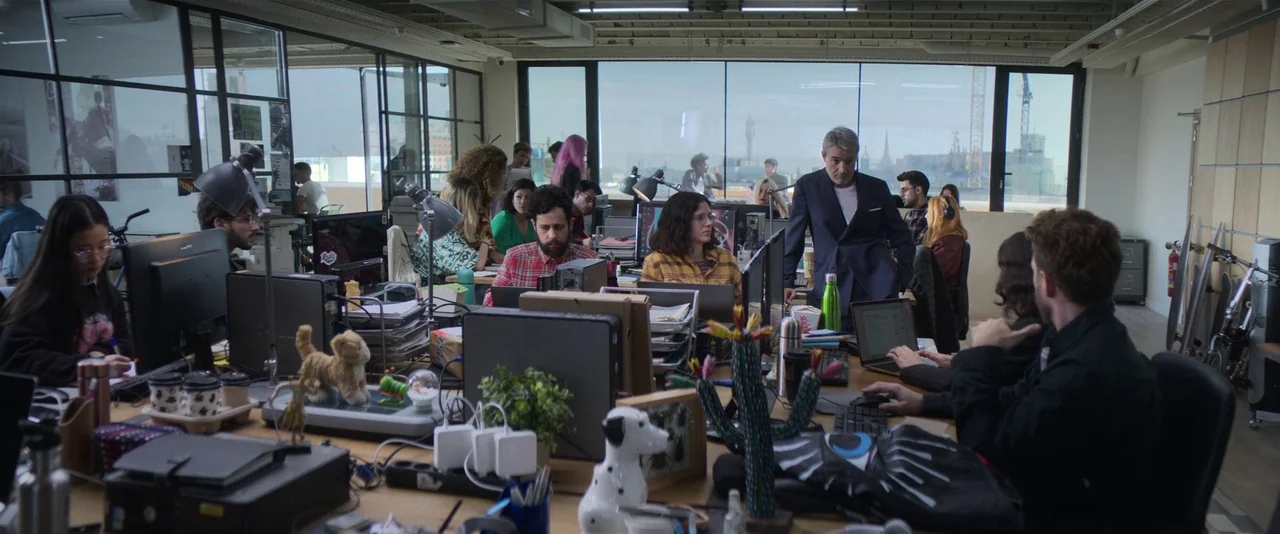 Crazy About Her (2021) movie still: wide — A busy open-plan office features long desks filled with computers and various personal items. Several…