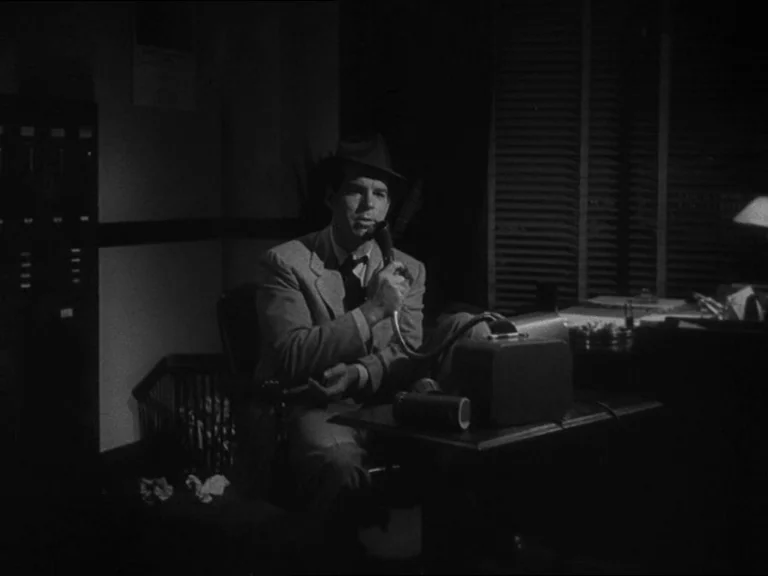 Double Indemnity (1944) movie still: medium — A dimly lit room contains a desk holding a dictation machine and paper records. A man sits at the…