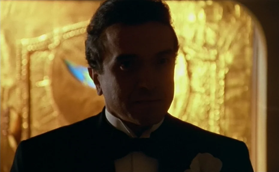 Life Is A Bed of Roses (1983) movie still: closeup — A dimly lit interior space features a warm glowing ornate gold background panel. A man dressed…