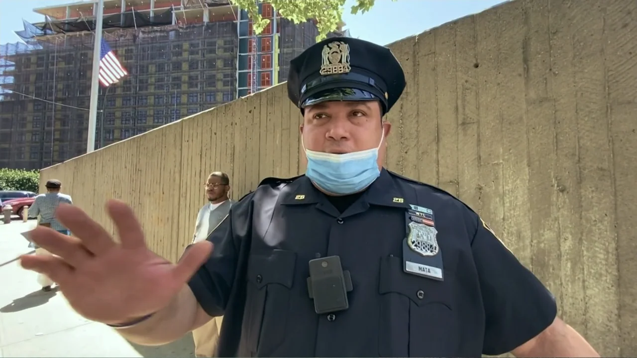 Covid Diaries NYC (2021) movie still: medium — A sidewalk sits adjacent to a tall concrete wall with a construction site beyond. An officer in…