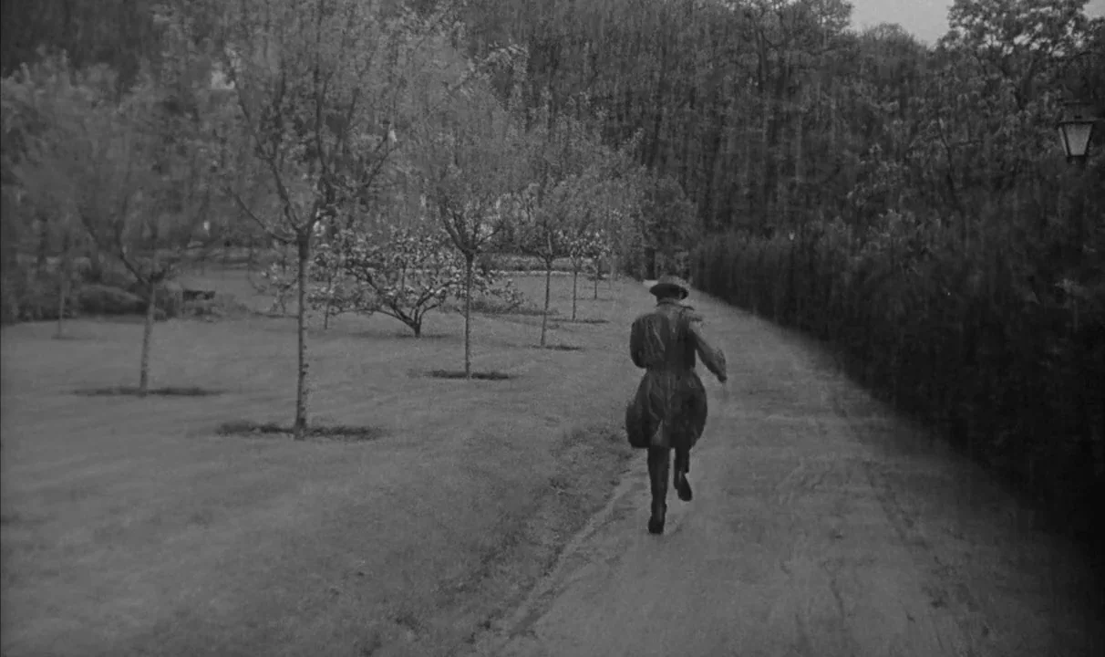 Le Doulos (1962) movie still: wide — A dirt path runs through a park lined with sparse trees and manicured hedges. A man in a long coat and hat runs…