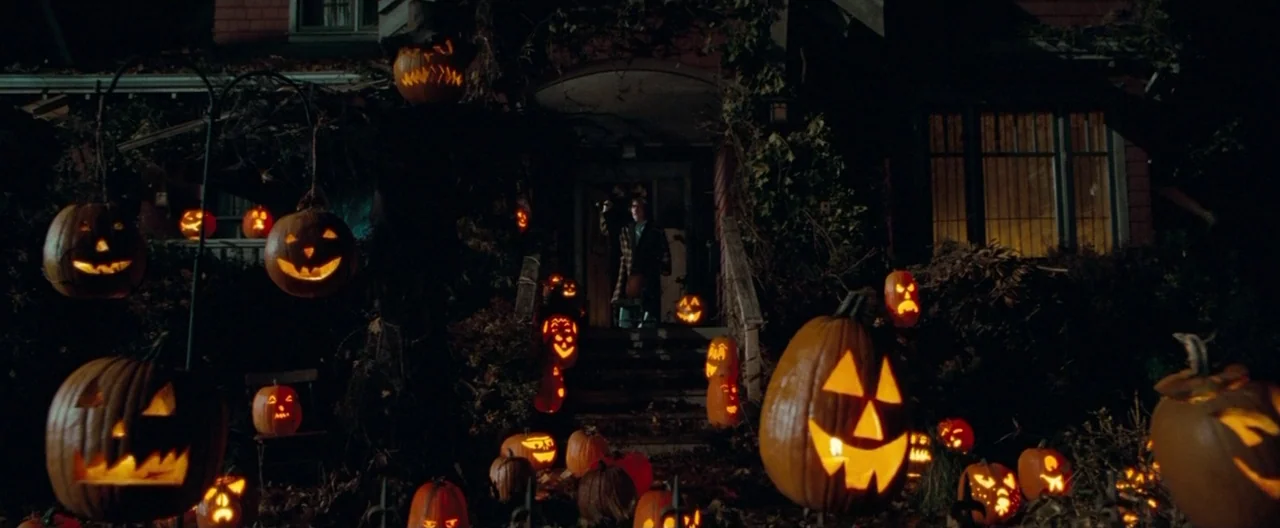 Trick ‘R Treat (2007) movie still: wide — A residential front porch and yard decorated with many glowing, carved jack-o'-lanterns at night. The…