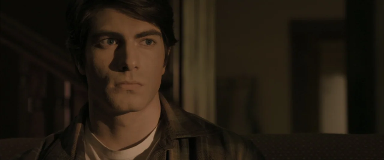 Superman Returns (2006) movie still: closeup — A dimly lit indoor space with blurred furniture in the background. A young man sits in the foreground…
