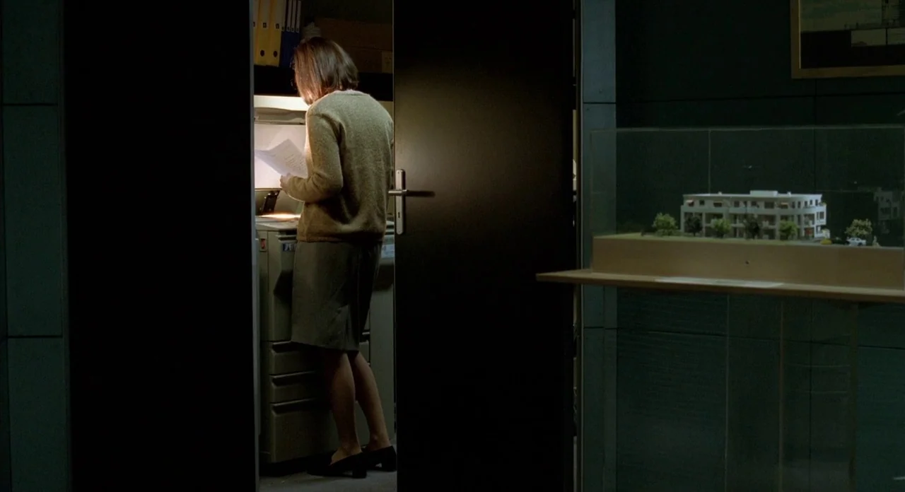 Read My Lips (2001) movie still: fullbody — A dimly lit room contains a photocopier and an architectural model on a display stand. A woman standing…