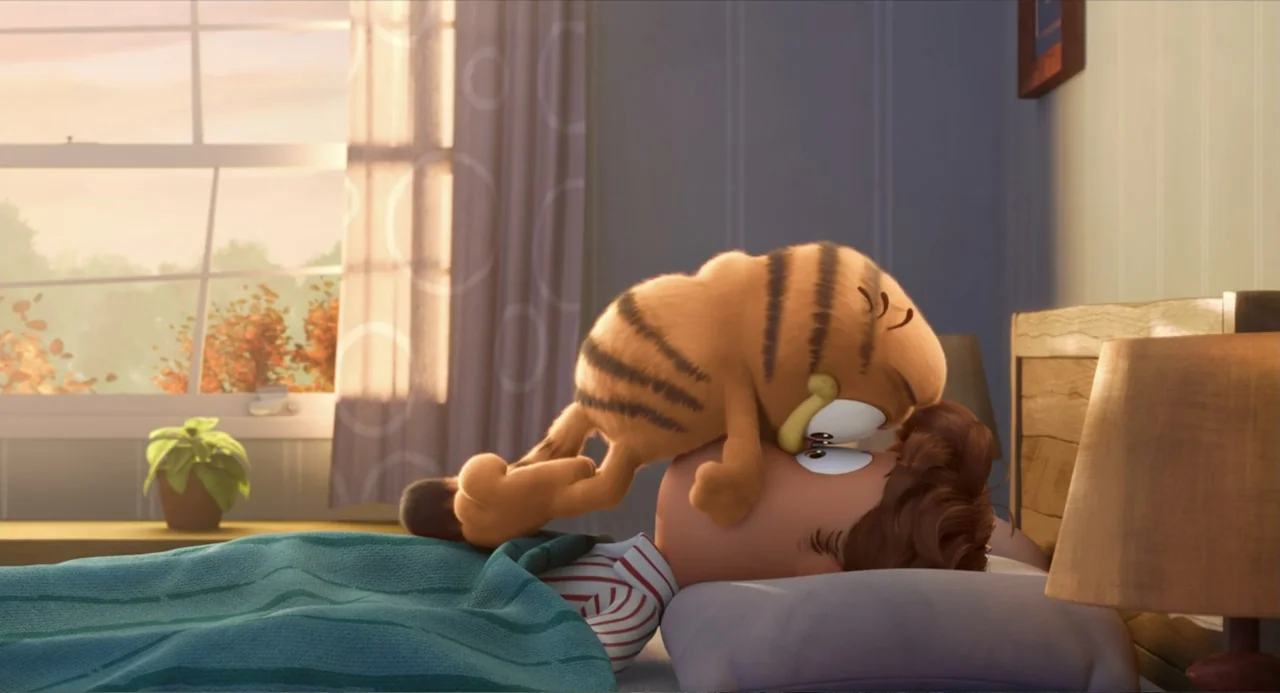 The Garfield Movie (2024) movie still: medium — A sunlit bedroom features a man sleeping in bed with a curtained window behind him. An orange cat…