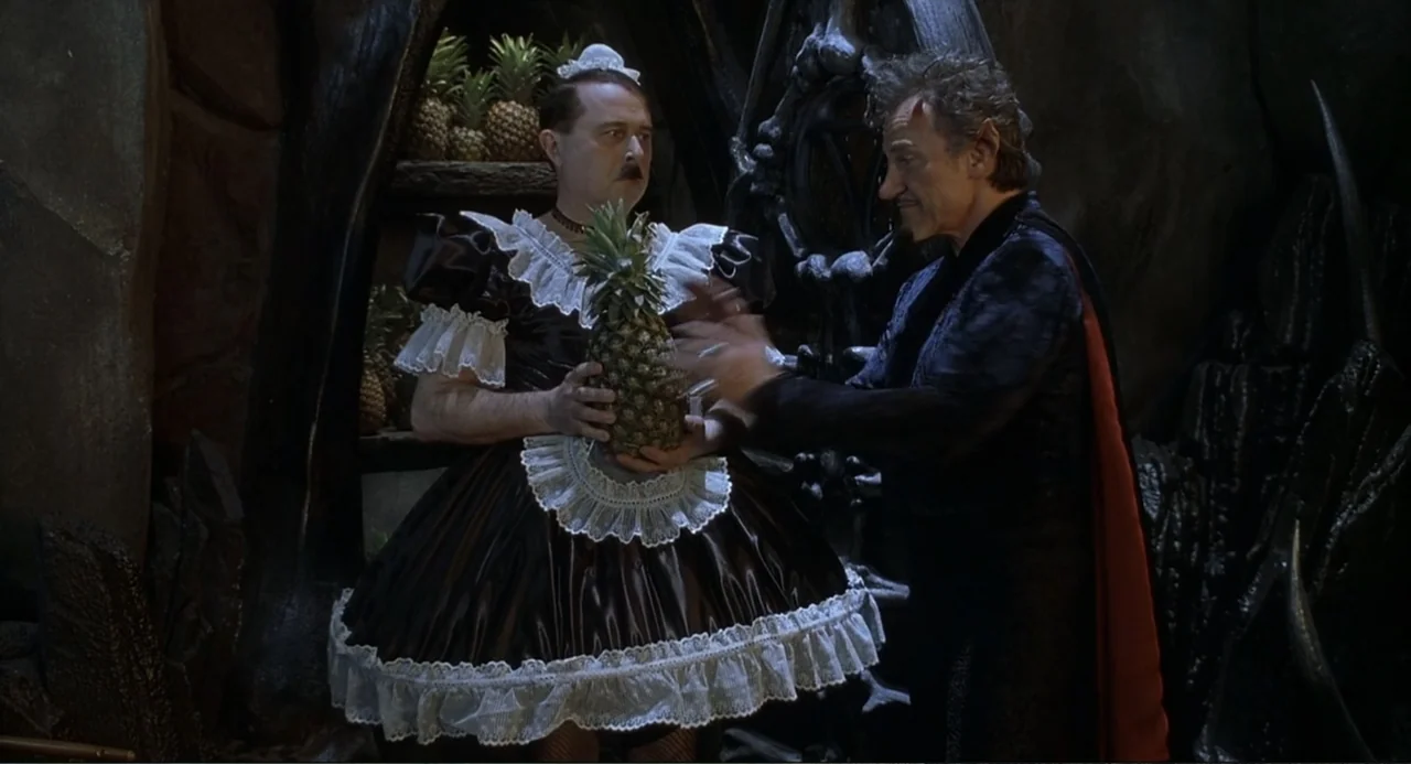 Little Nicky (2000) movie still: medium — A dark, rocky chamber contains shelves lined with several whole pineapples. A man in a maid costume stands…