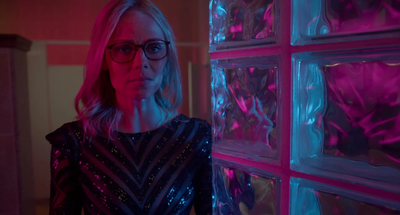 Rabid (2019) movie still: medium — A dimly lit washroom features a vertical wall of translucent glass blocks. A woman wearing glasses and a sequined…