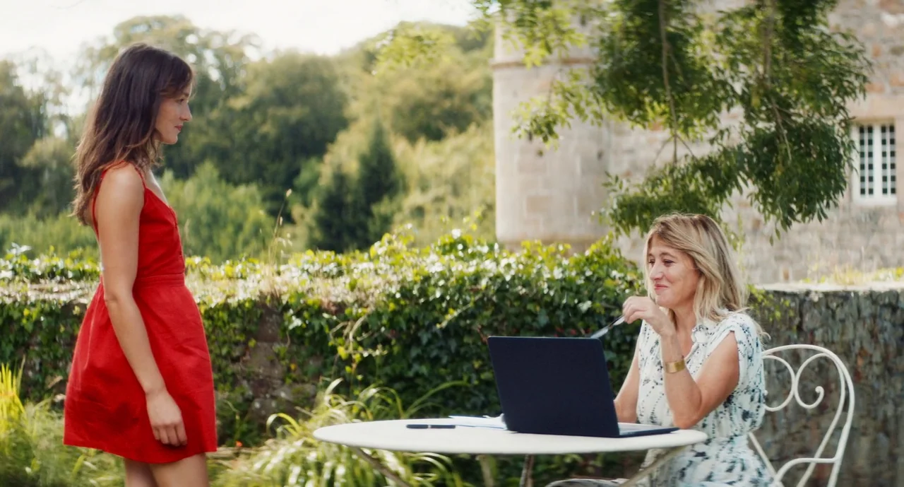 Anaïs in Love (2021) movie still: medium — An outdoor terrace overlooks a stone building surrounded by lush green trees and foliage. One woman in a…