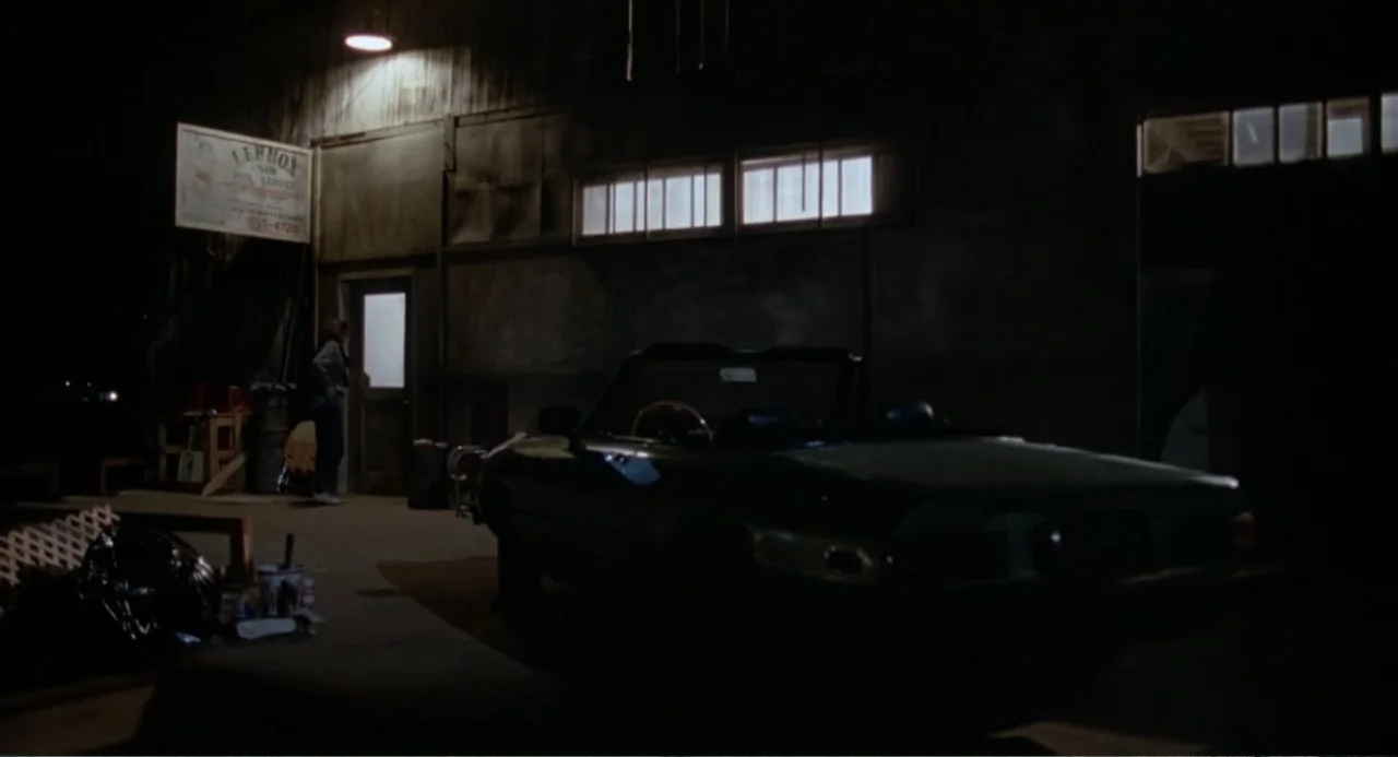 Dangerously Close (1986) movie still: wide — A dark exterior area beside a building at night with a parked convertible car. A single person stands…