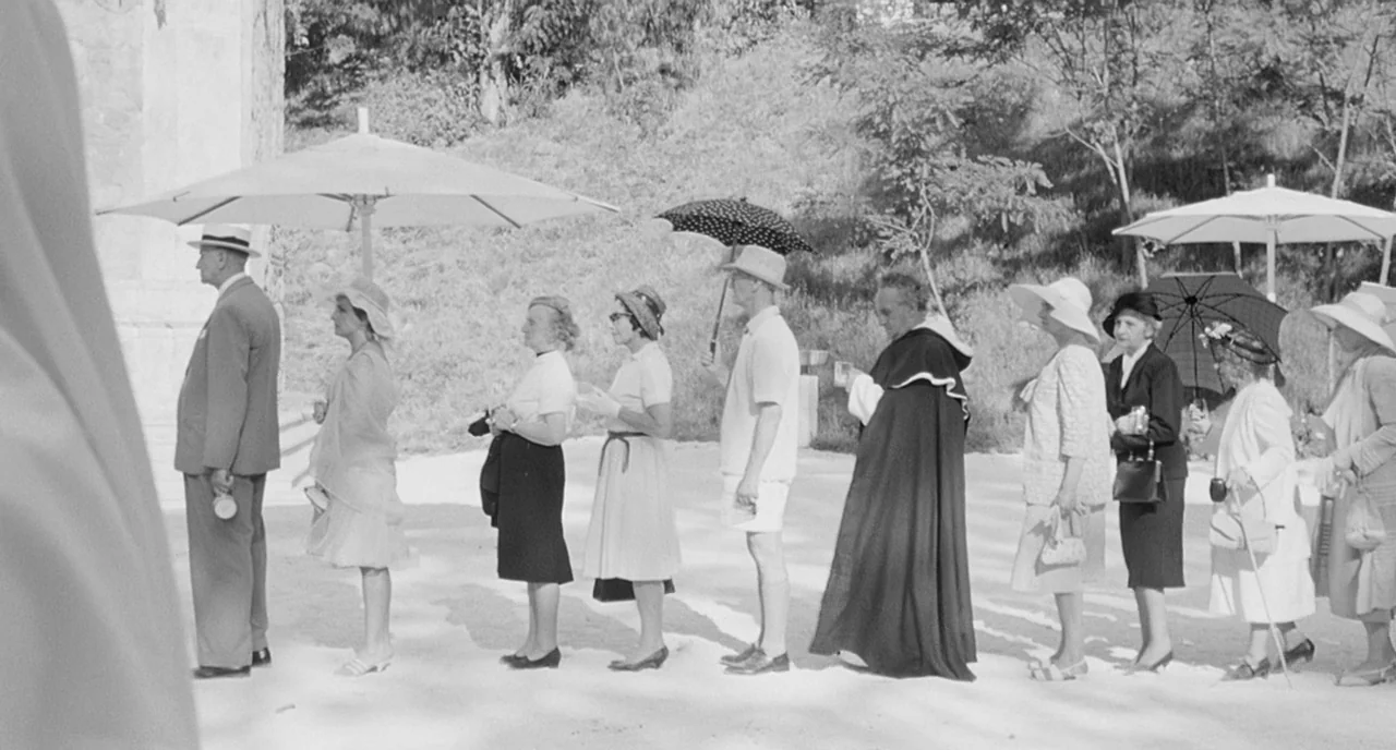 8 1/2 (1963) movie still: wide — A line of people in mid-20th century formal clothing stands on a gravel path. They wait in single file under…