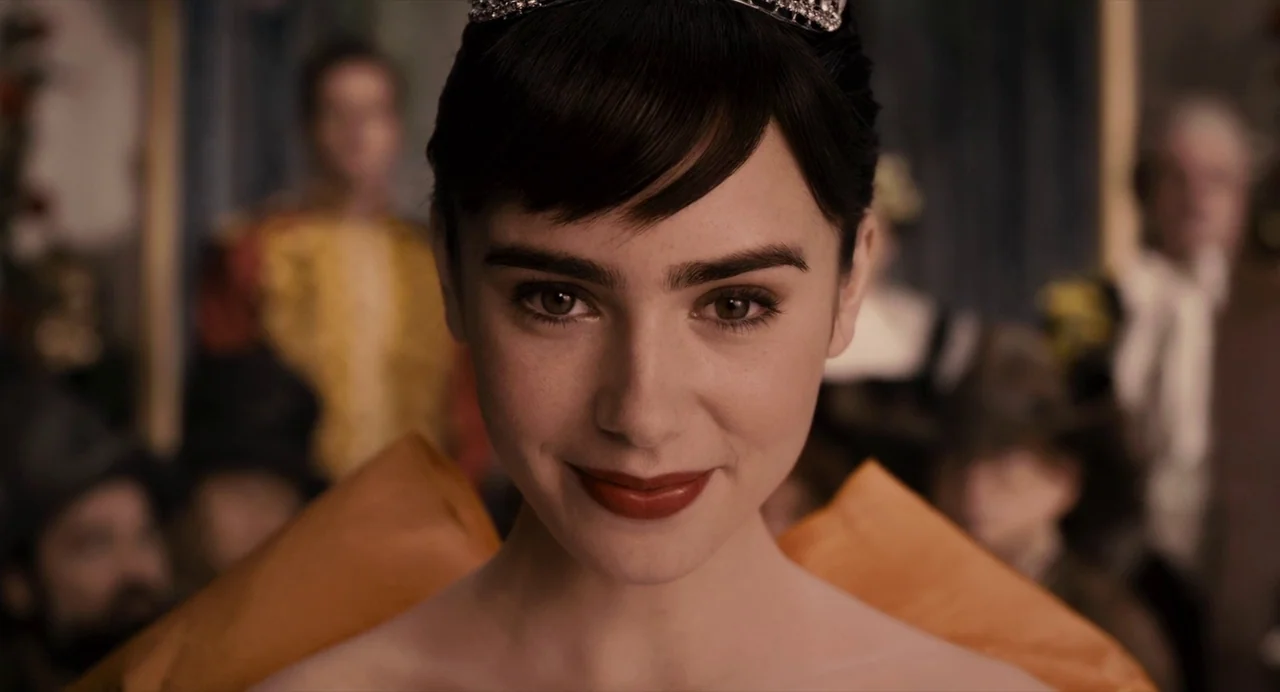 Mirror Mirror (2012) movie still: closeup — An indoor ballroom setting with blurred figures in formal attire in the background. A young woman wears…