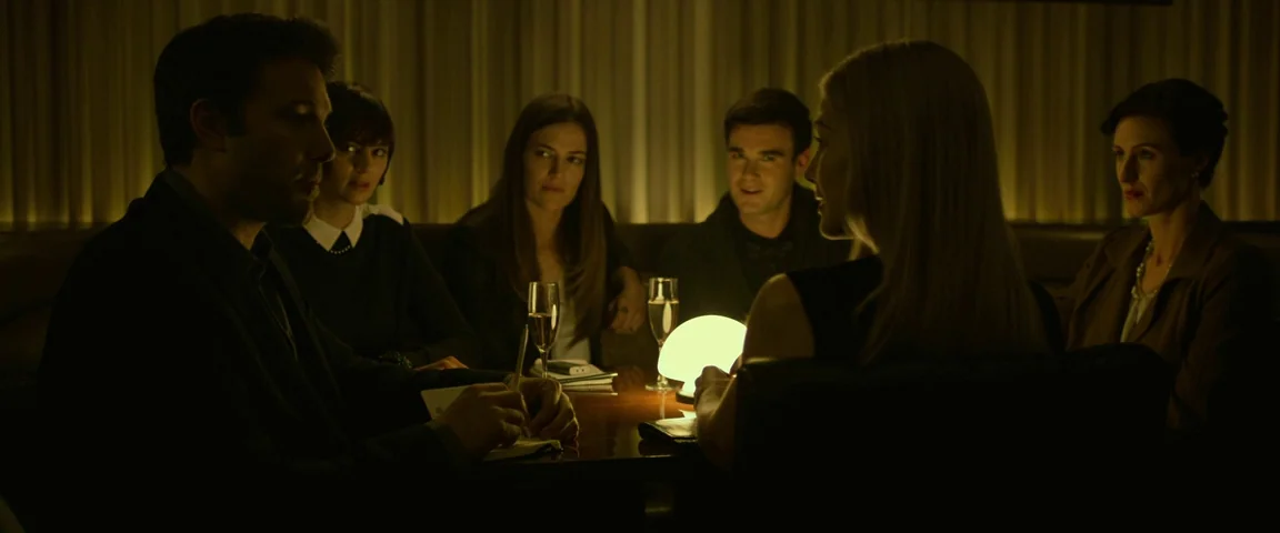 Gone Girl (2014) movie still: medium — A dimly lit restaurant booth features six people seated around a dark wooden table. A glowing modern lamp…