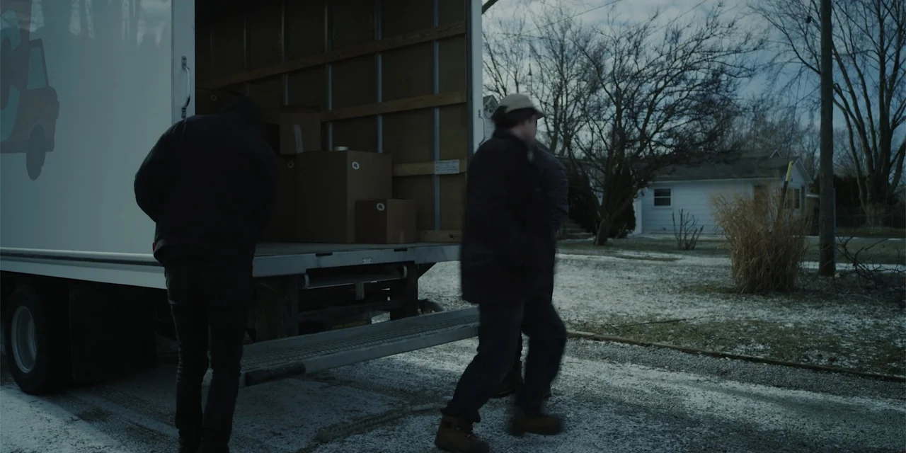 Lobos (2019) movie still: medium — A residential driveway with a large moving truck parked under a cloudy sky. Two men walk past the open rear of…
