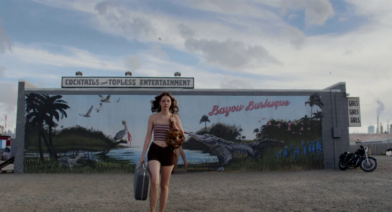 X (2022) movie still: fullbody — A gravel parking lot faces a large mural-painted wall advertising a burlesque venue. A young woman walks toward the…