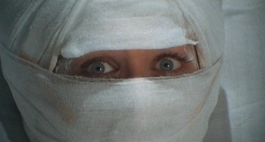 Blood From The Mummy’s Tomb (1971) movie still: closeup — A person lies on a clinical surface with their head wrapped completely in white bandages.…