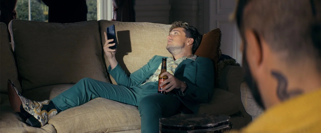 The Beekeeper (2024) movie still: medium — A man sits on a beige sofa in a room with light-colored walls. He holds a smartphone in one hand and a…