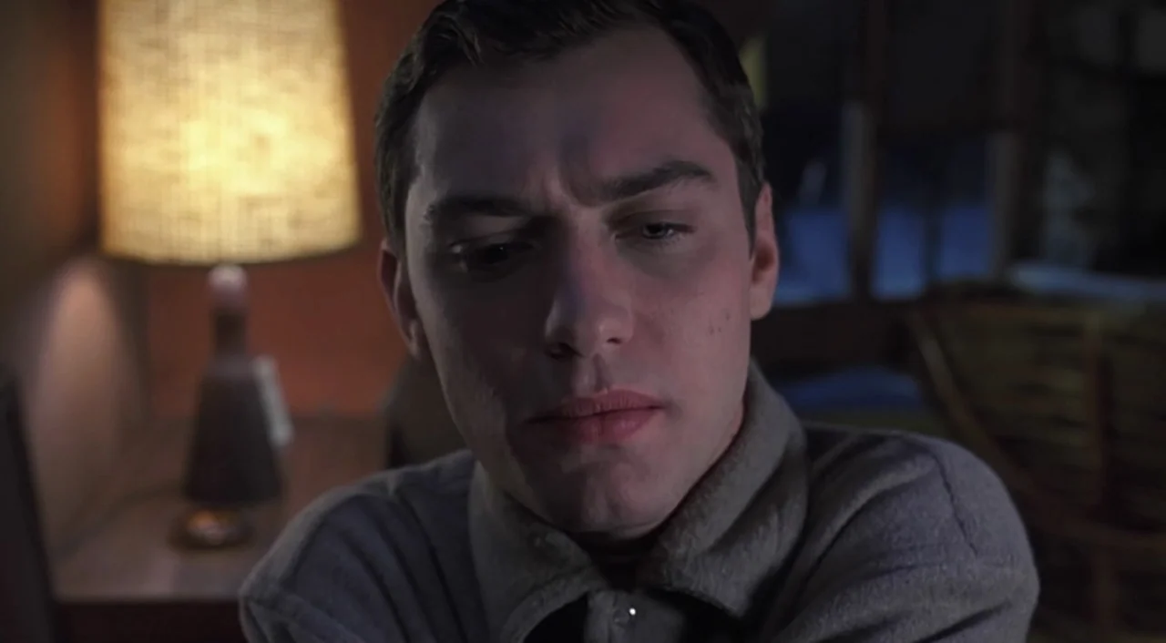 eXistenZ (1999) movie still: closeup — A dimly lit room with a bedside lamp providing a warm glow in back. A young man with dark hair sits in the…