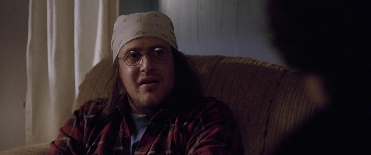 The End of the Tour (2015) movie still: medium — A man sits on a brown textured sofa in a dimly lit room. He wears a bandana and glasses while…