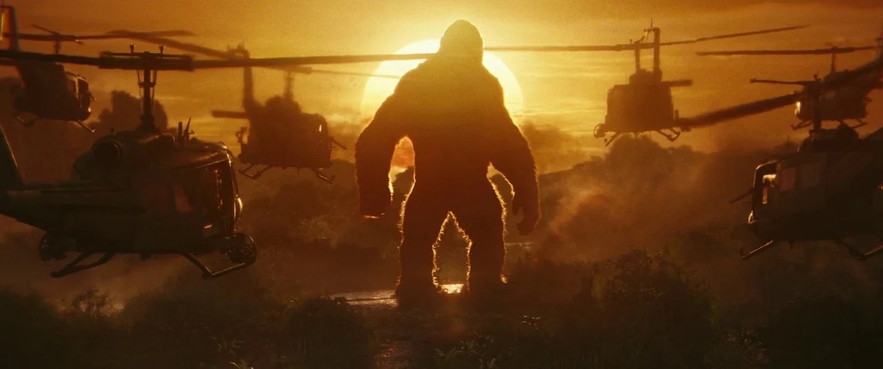 Kong: Skull Island (2017) movie still: wide — A giant gorilla stands in a tropical jungle clearing at sunset. Multiple military helicopters fly…