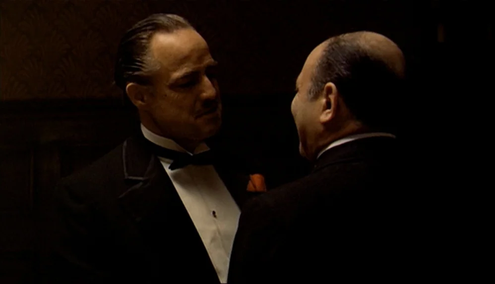 The Godfather (1972) movie still: medium — A dark, wood-paneled room serves as the setting for this quiet exchange. Two men stand in near-darkness…