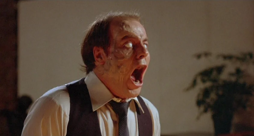 Scanners (1981) movie still: medium — A dimly lit interior room features a white wall and a blurred houseplant. A man in formal business attire…
