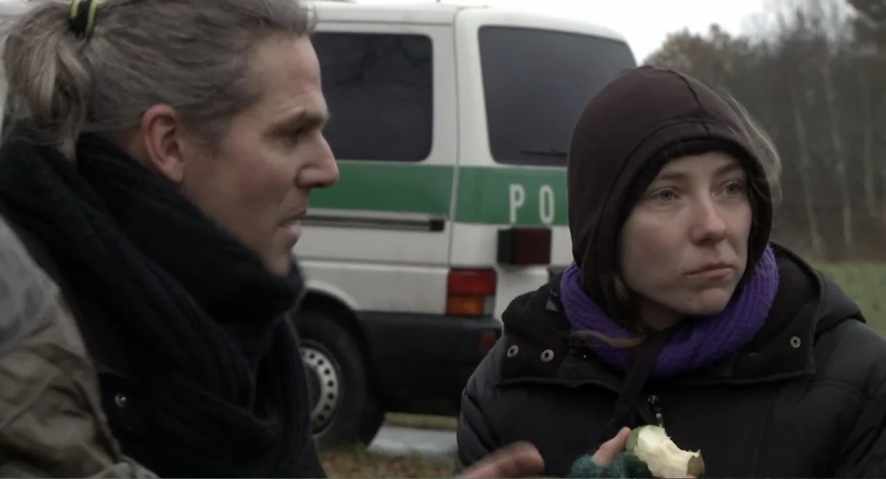 Az X- Kryptonit projekt (2020) movie still: medium — A woman in a hood and a man stand outside near a white police van. The woman holds a half-eaten…