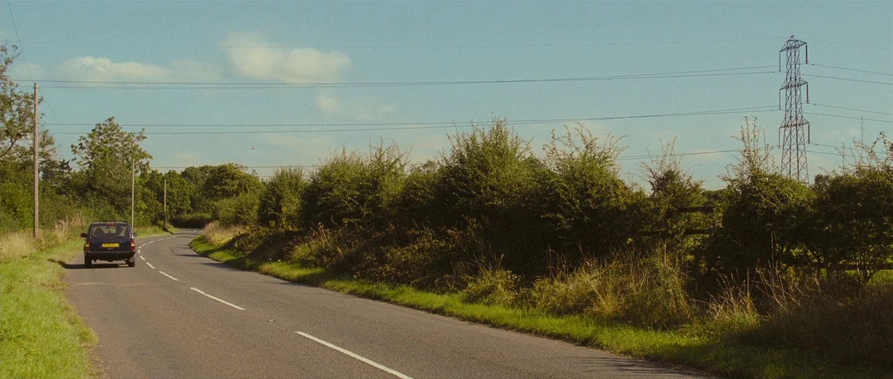 Eden Lake (2008) movie still: wide — A quiet asphalt road curves through a rural area lined with dense green bushes. High-voltage power lines extend…