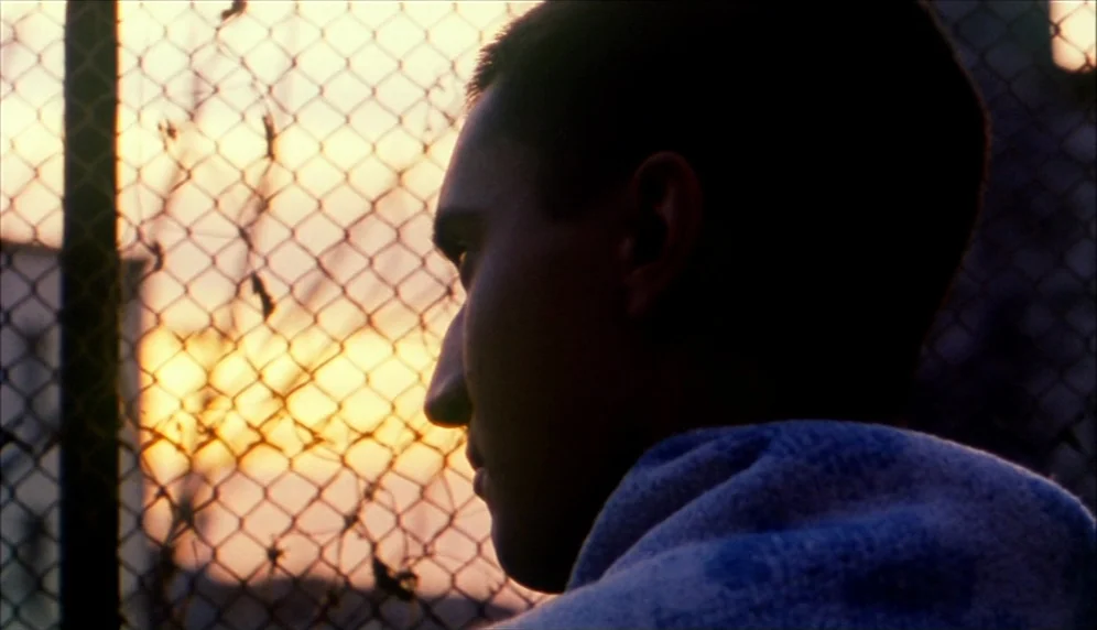 Nenette et Boni (1996) movie still: closeup — A young man is positioned in front of a chain link fence at dusk. The sunset provides a glowing yellow…