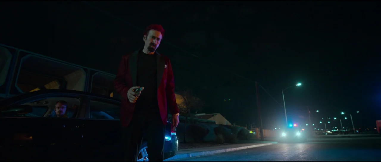 Sympathy For The Devil (2023) movie still: medium — An outdoor parking lot at night with a parked vehicle under distant streetlamps. A man with red…
