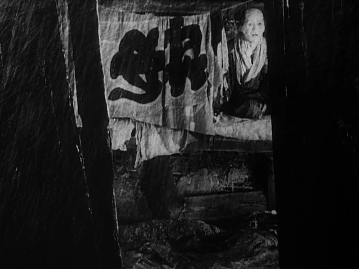 The Lower Depths (1957) movie still: medium — A dimly lit interior with low ceilings and draped fabric partitioning the space. An elderly man sits…