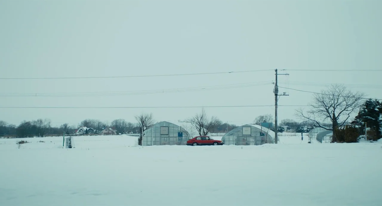 Drive My Car (2021) movie still: wide — A vast, snow-covered rural field features several greenhouse structures under an overcast, pale sky. A…