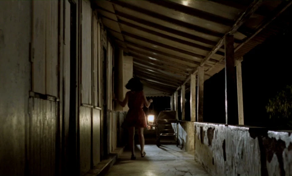 Casa De Lava (1994) movie still: fullbody — A weathered wooden veranda at night shows dark walls and a crumbling concrete railing. A young woman…