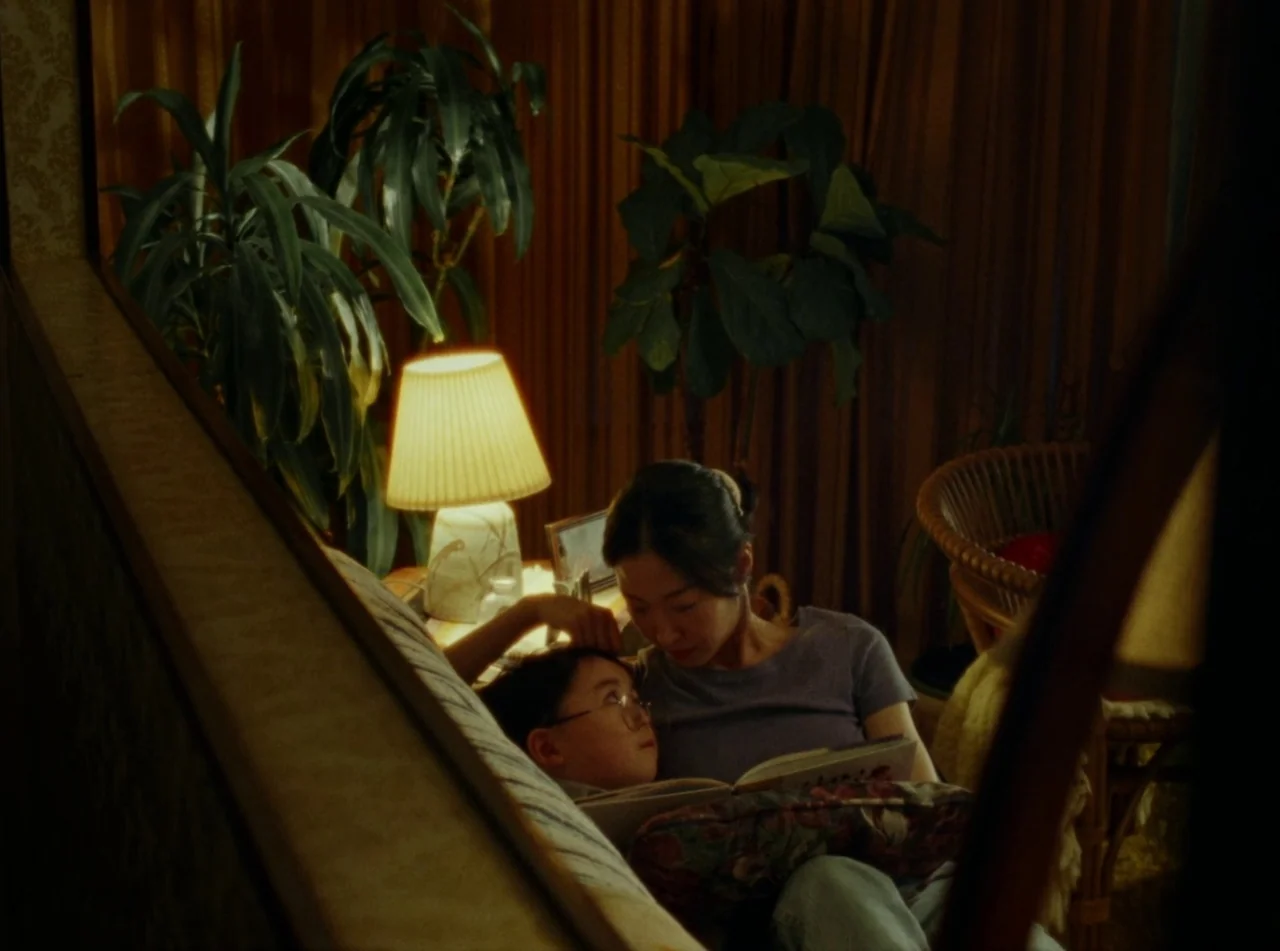 Riceboy Sleeps (2022) movie still: medium — A dimly lit room features dark curtains, lush indoor potted plants, and a wicker chair. A woman sits on…