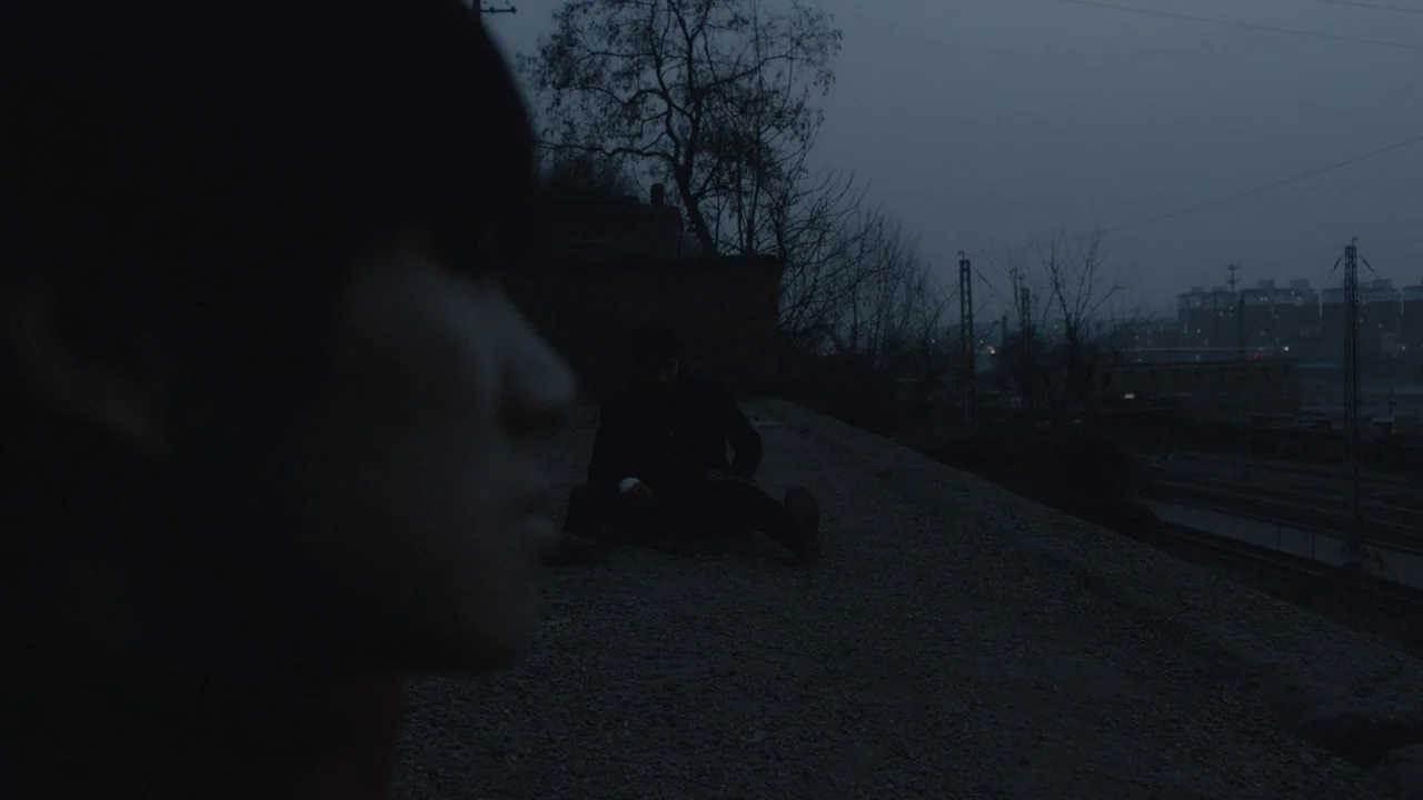 An Elephant Sitting Still (2018) movie still: wide — A dark, gravel-covered overpass surface during the dim light of blue hour. One man sits on the…