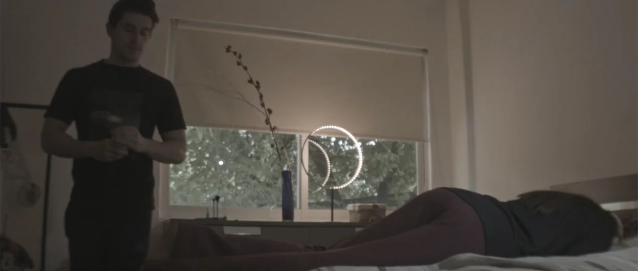 Amor Mutante (2019) movie still: medium — A spare, modern bedroom features a bed placed beneath a window with pulled blinds. A man stands near the…