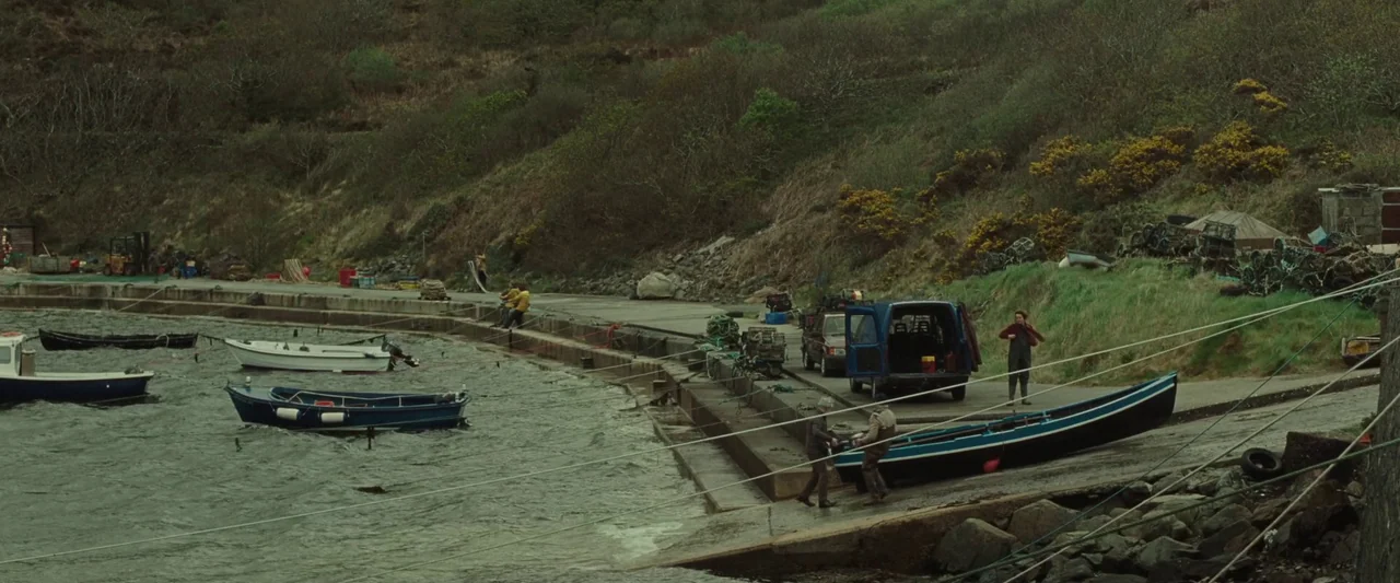 God’s Creatures (2022) movie still: wide — A remote concrete boat slip sits adjacent to a steep, vegetation-covered coastal hillside. Two men push a…
