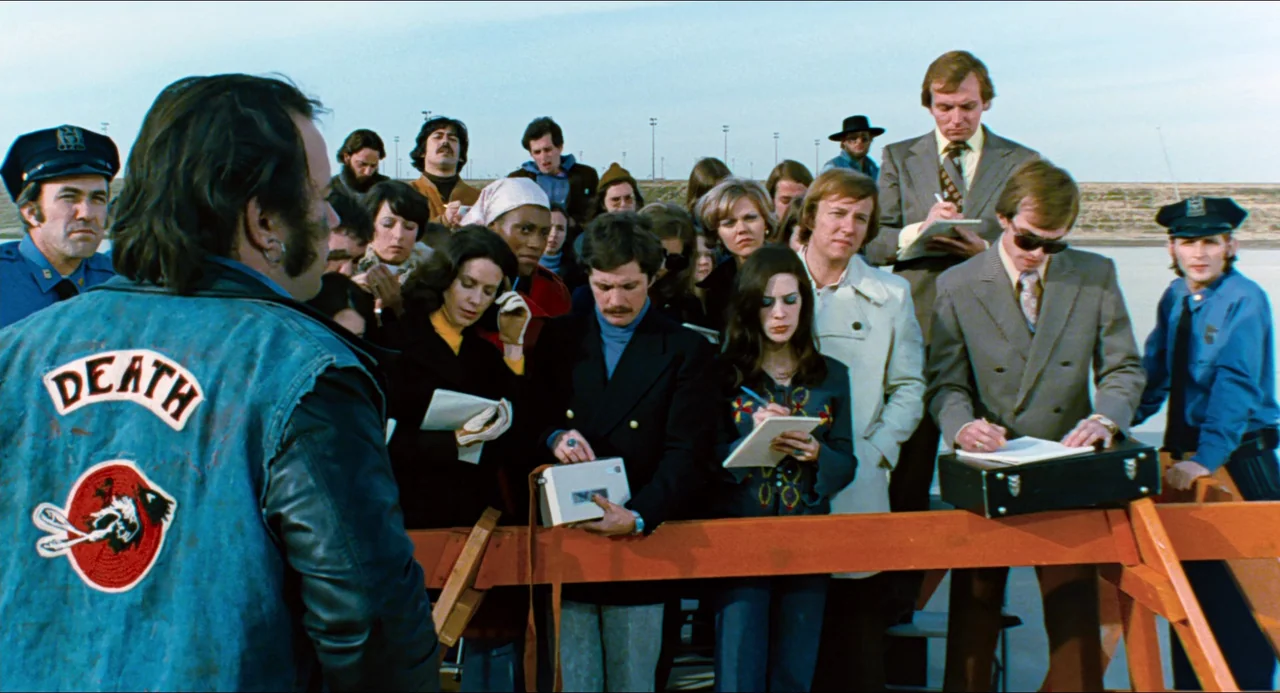 Phantom of the Paradise (1974) movie still: medium — A group of diverse reporters gather behind a wooden barricade holding notebooks and recorders.…