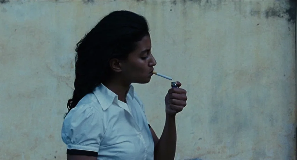Almayer’s Folly (2011) movie still: medium — A woman stands in profile against a plain, textured, light-colored concrete wall. She holds a cigarette…