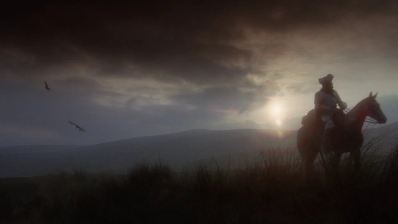 Excalibur (1981) movie still: wide — A vast, open moorland sits under a dark, heavy sky at sunset. A lone rider on horseback pauses on the grass as…